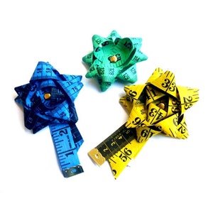 Könnte beinhalten: Drei bunte Schleifen aus Maßband. Die Schleifen sind blau, grün und gelb. Die blaue Schleife hat ein Maßband daran befestigt.