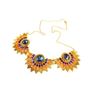 Könnte beinhalten: Eine goldene Kettenkette mit drei Perlen-Sonnenstrahl-Anhängern. Jeder Anhänger hat einen großen blauen Edelstein, der von kleineren violetten und orangefarbenen Perlen umgeben ist.