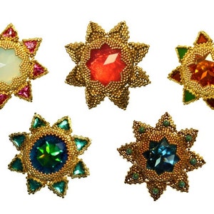 Puede incluir: Cinco broches de oro en forma de estrella con piedras preciosas de diferentes colores en el centro. Las piedras preciosas son rojas, naranjas, verdes, azules y blancas.