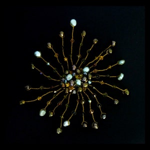 Pode incluir: Um pente de cabelo de fio dourado com um grupo de contas azuis iridescentes e marrons no centro. As contas são dispostas em um padrão circular e são cercadas por contas douradas menores.