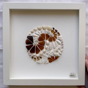 May include: Framed artwork with a circular design made from seashells and sea glass. The artwork is set against a white canvas within a white frame. The sea glass is brown and the seashells are white.