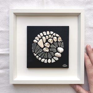 Peut inclure: Une œuvre d'art encadrée présentant un arrangement circulaire de verre de mer blanc, de coquillages et de pierres sur un fond noir. L'œuvre est encadrée dans un cadre carré blanc, créant une esthétique minimaliste. La composition est un design unique et naturaliste.