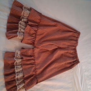 May include: A pair of bloomers featuring a red and white vertical striped pattern. The garment has an elasticized waistband and wide, ruffled leg openings that are trimmed with a delicate, off-white floral lace border along the edges.