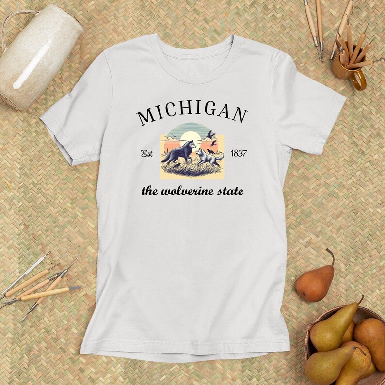 Pode incluir: Camiseta branca com um design gr&aacute;fico preto que diz "MICHIGAN" com uma cena de p&ocirc;r do sol e dois lobos. O texto abaixo do gr&aacute;fico diz "Est. 1837 the wolverine state".