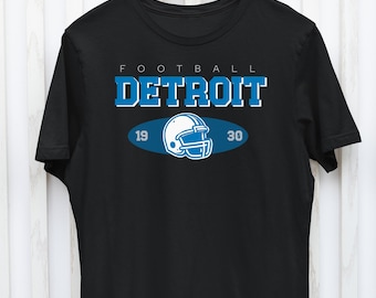 Op de afbeelding: Zwart T-shirt met een blauwe en witte voetbalhelm afbeelding en de tekst "FOOTBALL DETROIT 19 30".