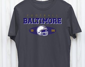 Puede incluir: Camiseta gris oscuro con temática de fútbol. La camiseta presenta la palabra "FOOTBALL" en dorado y "BALTIMORE" en morado. Debajo hay un gráfico de un casco de fútbol blanco dentro de un óvalo azul, con "19" y "96" a cada lado.