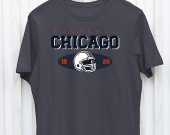 May include: Dark gray t-shirt with the word "FOOTBALL" in orange, and "CHICAGO" in white with a navy blue outline. Below is a football helmet graphic and the numbers "19" and "20". The shirt is hanging on a wooden hanger.