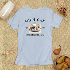 Pode incluir: Camiseta azul claro com um design gr&aacute;fico preto que apresenta uma cena de p&ocirc;r do sol com dois lobos e o texto "MICHIGAN Est 1837 the wolverine state".