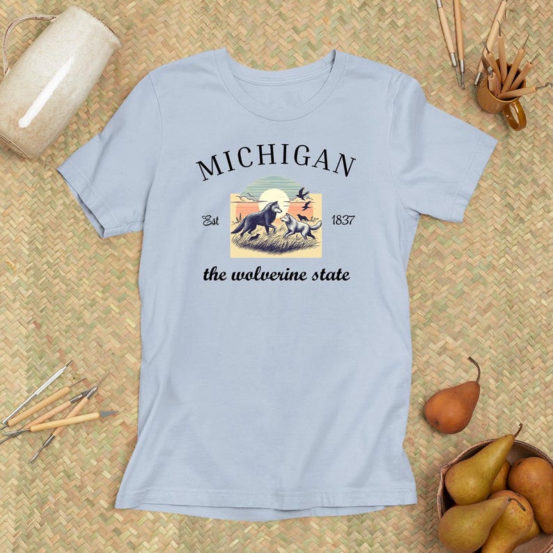 Pode incluir: Camiseta azul claro com um design gr&aacute;fico preto que apresenta uma cena de p&ocirc;r do sol com dois lobos e o texto "MICHIGAN Est 1837 the wolverine state".