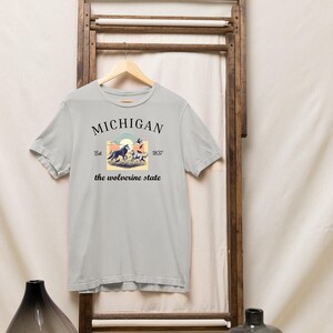 Pode incluir: Uma camiseta cinza claro com um design gr&aacute;fico de estilo vintage que apresenta um lobo e o texto "MICHIGAN Est. 1837 the wolverine state".