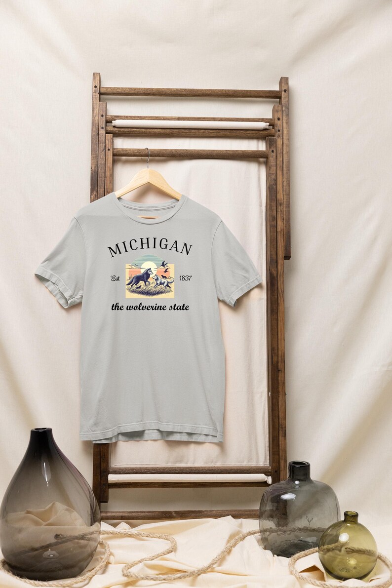 Pode incluir: Uma camiseta cinza claro com um design gr&aacute;fico de estilo vintage que apresenta um lobo e o texto "MICHIGAN Est. 1837 the wolverine state".
