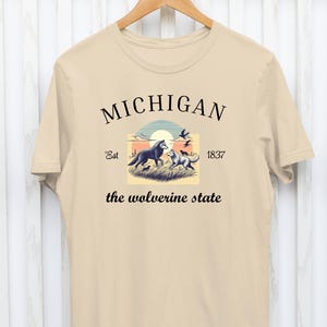 Pode incluir: Uma camiseta bege com um design gr&aacute;fico que apresenta uma cena de p&ocirc;r do sol com dois lobos e o texto "MICHIGAN Est 1837 the wolverine state".