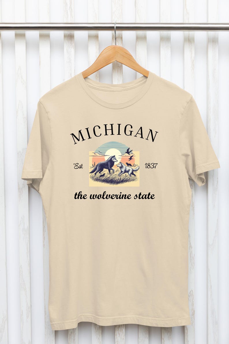 Pode incluir: Uma camiseta bege com um design gr&aacute;fico que apresenta uma cena de p&ocirc;r do sol com dois lobos e o texto "MICHIGAN Est 1837 the wolverine state".