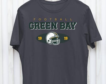 Puede incluir: Una camiseta gris oscuro con el texto "FOOTBALL GREEN BAY" en letras verdes y amarillas. Un casco de fútbol está centrado debajo del texto con los números "19" a cada lado.