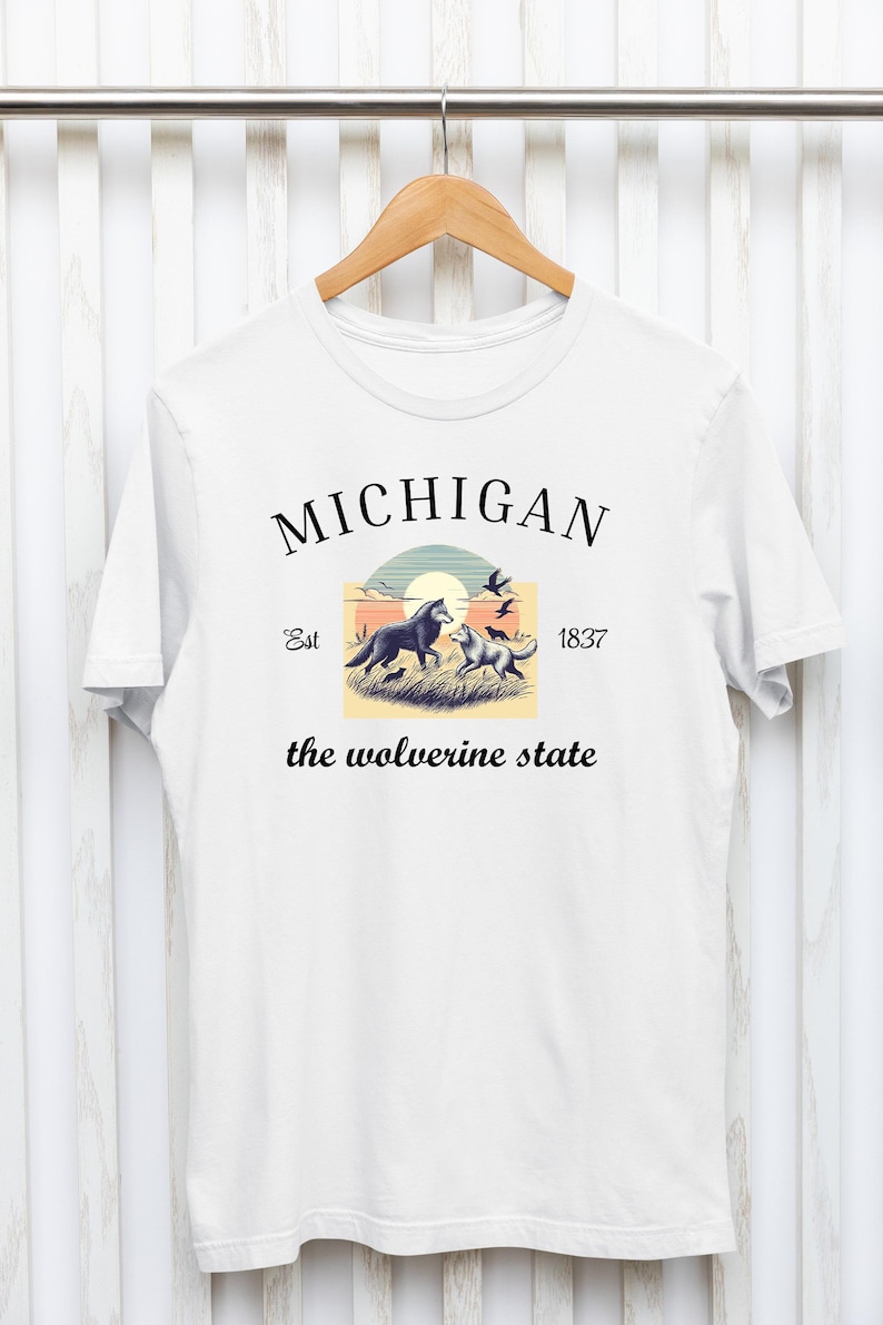 Pode incluir: Camiseta branca com um design gr&aacute;fico que apresenta uma cena de p&ocirc;r do sol com dois lobos e p&aacute;ssaros voando acima. O texto "MICHIGAN Est 1837 the wolverine state" &eacute; impresso na camiseta.