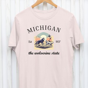 Pode incluir: Uma camiseta rosa com um design gr&aacute;fico que apresenta um p&ocirc;r do sol, dois lobos e o texto "MICHIGAN Est 1837 the wolverine state".