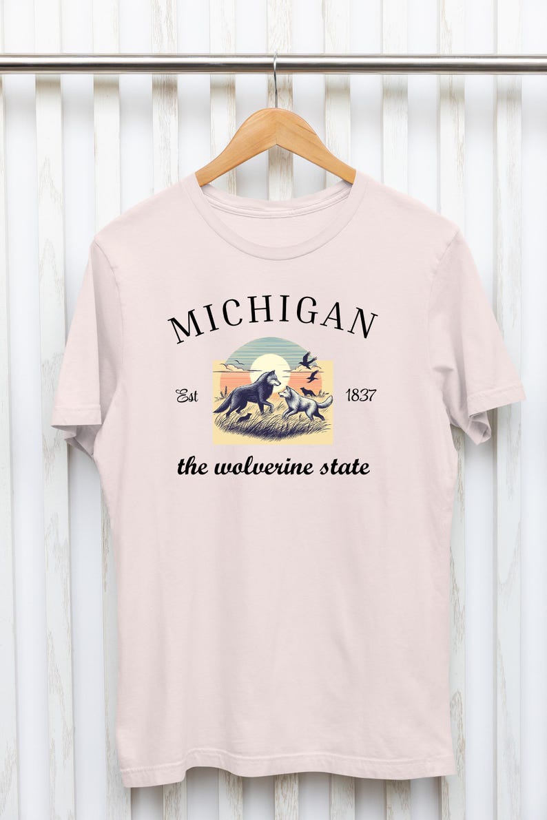 Pode incluir: Uma camiseta rosa com um design gr&aacute;fico que apresenta um p&ocirc;r do sol, dois lobos e o texto "MICHIGAN Est 1837 the wolverine state".