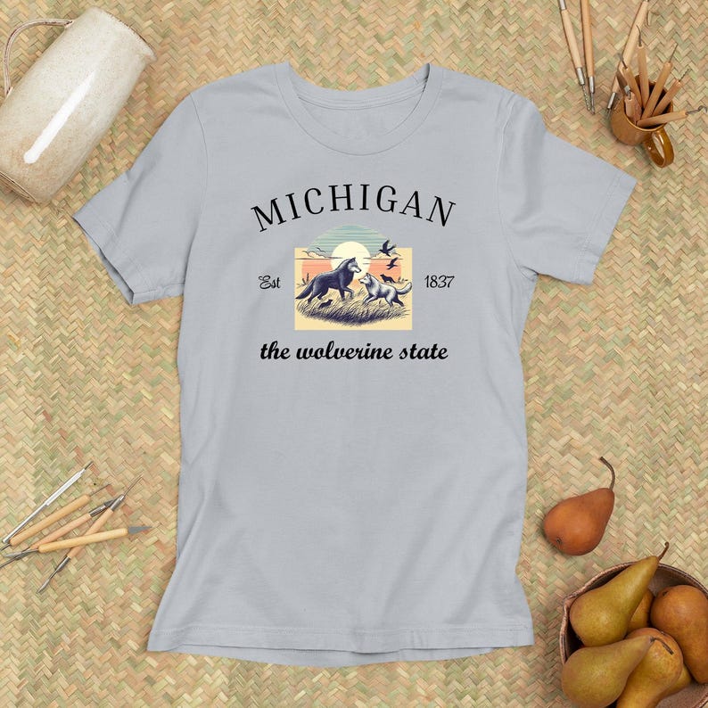 Pode incluir: Uma camiseta azul clara com um design gr&aacute;fico que apresenta uma cena de p&ocirc;r do sol com dois lobos e o texto "MICHIGAN Est 1837 the wolverine state".