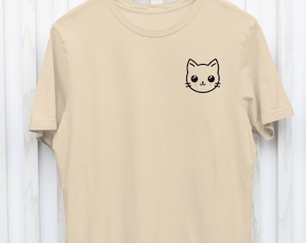 May include: A beige t-shirt with a small, black cartoon cat face on the upper left chest. The shirt is hanging on a wooden hanger against a white and beige striped background. The cat face has large eyes and a small nose.