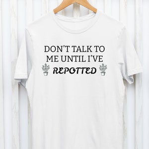 Könnte beinhalten: Weißes T-Shirt mit dem Text "Sprich mich nicht an, bevor ich nicht umgetopft habe" und einer kleinen Topfpflanzengrafik auf jeder Seite des Textes.