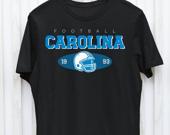 Może przedstawiać: Czarny T-shirt z niebiesko-białym nadrukiem przedstawiającym kask do futbolu amerykańskiego i tekst "FOOTBALL CAROLINA 19 93".