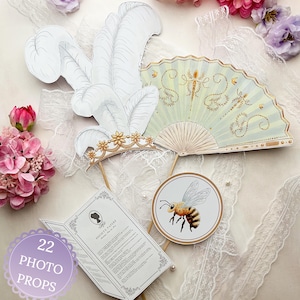 May include: A collection of 22 photo props, including a white feather headdress, a light green fan with gold accents, a round bee illustration, and a booklet labeled "Society Papers". The props are arranged on a white lace surface.