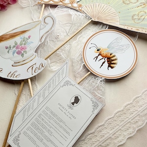 K&ouml;nnte beinhalten: Verschiedene dekorative Papierausstiche auf Holzst&auml;ben, darunter eine Teetasse mit Blumendesign, eine Biene, ein F&auml;cher und ein Dokument mit der Aufschrift "Society Papers". Die Artikel sind in Pastellt&ouml;nen mit goldenen Akzenten gehalten, was ein Vintage- oder viktorianisches Thema suggeriert.
