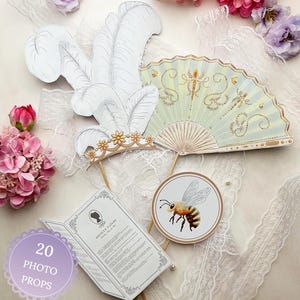 May include: A collection of photo props, including a white feather headdress with a gold crown, a pale green fan with gold accents, a round bee illustration, and a booklet. The props are arranged on a white lace surface. The text "20 PHOTO PROPS" is visible.