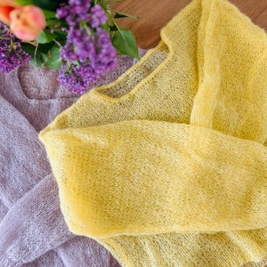 Puede incluir: Dos suéteres de punto transparente, en colores pastel suaves, uno lavanda y otro amarillo, se muestran sobre una superficie de madera. Un ramo de lilas moradas y tulipanes naranjas añade un toque primaveral. Los suéteres tienen un tejido abierto y delicado.