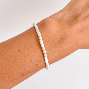 Op de afbeelding: Een delicate witte kralenarmband met kleine, ronde parels en langwerpige, rechthoekige kralen. De armband wordt om een pols gedragen en toont zijn eenvoudige maar elegante ontwerp. De kralen zijn aan elkaar geregen, waardoor een subtiel, minimalistisch accessoire ontstaat.