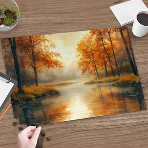 May include: A rectangular jigsaw puzzle featuring an autumn forest landscape with orange and yellow trees reflected in a calm river. The scene depicts a bright, hazy light filtering through the trees, with a hand placing a final puzzle piece.