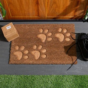 May include: A brown coir doormat with four paw prints embossed in the surface. The doormat is placed on a grey surface with green grass in the foreground.