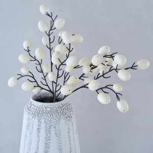 May include: A white ceramic vase with a textured surface holds a branch of white faux eggs with black stems.