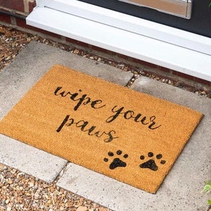 Könnte beinhalten: Eine braune Kokosmatte mit dem Text "wipe your paws" in schwarzer Schrift. Zwei schwarze Pfotenabdrücke befinden sich unter dem Text.