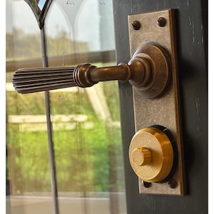 May include: A close-up of a brass door handle with a fluted design. The handle is attached to a rectangular plate with two screws. A gold-colored knob is visible below the handle.