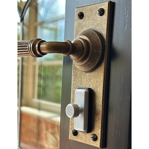 Op de afbeelding: Close-up van een vintage deurklink en slotset. De hendel en achterplaat hebben een bronzen afwerking, met een gedetailleerd, gestructureerd ontwerp. Het vergrendelingsmechanisme is wit en grijs, gemonteerd op een donkere houten deur.