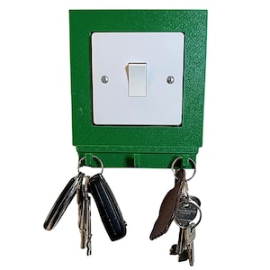 Könnte beinhalten: Ein grüner, wandmontierter Schlüsselhalter mit einer weißen Lichtschalterplatte und zwei Schlüsselhaken. Der Schlüsselhalter hat ein dekoratives Design um die Schalterplatte herum. An den Haken hängen Schlüssel.