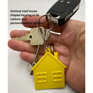 Puede incluir: Un llavero amarillo con forma de casa con una cadena y un anillo plateados. El llavero tiene dos ventanas y una chimenea. El llavero está unido a un juego de llaves.