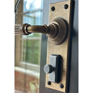 May include: A brass door handle with a decorative design and a black push button. The handle is mounted on a brass plate with screws.