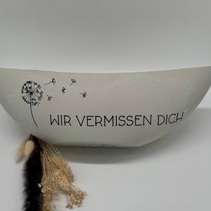 Könnte beinhalten: Eine weiße, ovale Keramikschale mit einer schwarzen Pusteblumen-Illustration und dem deutschen Text "WIR VERMISSEN DICH". Die Schale hat eine glatte Oberfläche und ein schlichtes, elegantes Design. Getrocknete Blumen und dekorative Elemente sind darunter sichtbar.