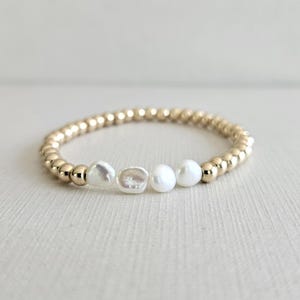 May include: A gold beaded bracelet with three white pearls. The bracelet features small, round gold beads and three irregularly shaped pearls. The pearls are centered on the bracelet, with the gold beads on either side.