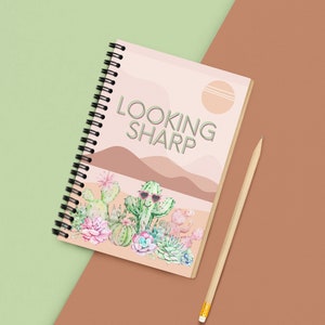 Peut inclure: Cahier à spirales avec une couverture représentant un paysage désertique et le texte "LOOKING SHARP". Le motif comprend des cactus, des plantes succulentes et un cactus portant des lunettes de soleil. Un crayon en bois est posé à côté du cahier.