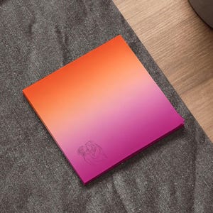 Könnte beinhalten: Ein quadratischer Notizblock mit einem Farbverlauf von Orange nach Rosa. Eine Strichzeichnung von zwei sich küssenden Personen befindet sich in der unteren rechten Ecke. Der Notizblock liegt auf einer grauen Oberfläche.