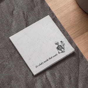 May include: A square, off-white notepad with a distressed texture. A black and white illustration of a skeleton holding a rose is in the corner. The text "it's what's inside that counts" is printed in black.