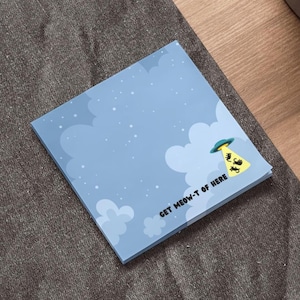 May include: A square notepad with a light blue background featuring white clouds and stars. A yellow beam shines from a UFO, illuminating black cat silhouettes. The text "GET MEOW-T OF HERE" is printed on the notepad.