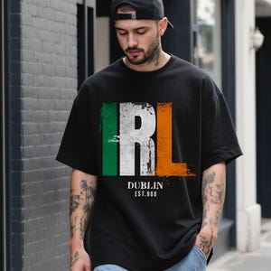 Op de afbeelding: Zwart T-shirt met de letters "IRL" in wit, met groene en oranje verticale strepen. De woorden "DUBLIN" en "EST.988" staan eronder. Het shirt heeft een casual stijl.