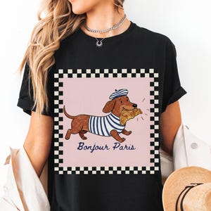 Op de afbeelding: Zwart T-shirt met een cartoon teckel met een gestreept shirt en baret, met een croissant. Het ontwerp staat in een roze vierkant met een zwart-witte geblokte rand. De tekst "Bonjour Paris" staat eronder.
