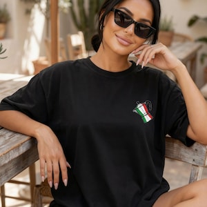 Op de afbeelding: Zwart T-shirt met een kleine geborduurde Italiaanse koffiezetter in de kleuren van de Italiaanse vlag. Het shirt heeft een relaxte pasvorm en wordt buiten gedragen. De afbeelding bevat ook een drankje in een doorzichtige plastic beker.