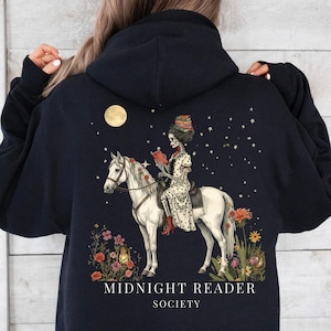 Può includere: Felpa con cappuccio blu navy con un design artistico. Raffigura uno scheletro a cavallo che legge un libro sotto la luna e le stelle. Il design include fiori e il testo "MIDNIGHT READER SOCIETY". Un capo unico.