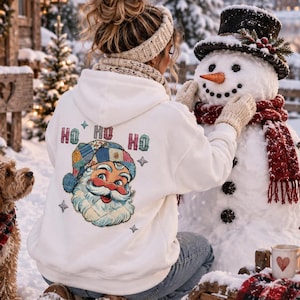 Op de afbeelding: Een witte hoodie met een afbeelding van een vintage-stijl kerstmangezicht met een patchworkmuts, omringd door de tekst HO HO HO en kleine sterpictogrammen. Het ontwerp is gedrukt op de achterkant van het kledingstuk dat buiten wordt gedragen.